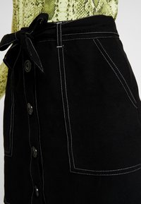 Černá sukně s předními knoflíky, bílým prošíváním, velkými kapsami a zavazovacími pásky na osobě, která má na sobě zelený vzorovaný top.