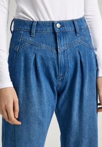 Pantalones de mezclilla azul con un frente plisado y una cintura alta. El diseño incluye pasadores para cinturón y costuras en naranja contrastante.