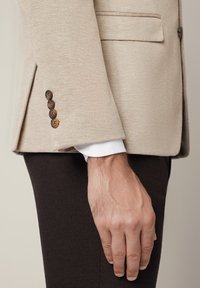 Beżowa teksturowana rękawka marynarki z czterema brązowymi guzikami, mankiet białej koszuli i zrelaksowana ręka odpoczywająca obok ciemno-brązowych spodni.