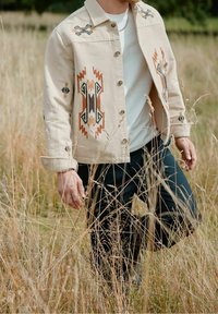 Beige gebloemde jas met borduursels in bruin, zwart en roestkleur. Zwarte broek, handen bezig met hoge grassprieten.