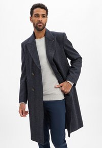 Donkerblauwe visgraatjas met een dubbelgeknoopte sluiting, gouden knopen en grote revers, gedragen over een lichtgrijze sweatshirt en donkerblauwe broek.