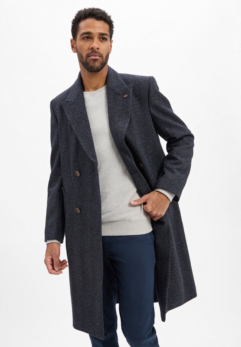 Donkerblauwe visgraatjas met een dubbelgeknoopte sluiting, gouden knopen en grote revers, gedragen over een lichtgrijze sweatshirt en donkerblauwe broek.