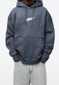 Šedá mikina s kapucí, přední kapsou a bílým logem. Materiál z bavlněné směsi, žebrované manžety a lem, uvolněný střih a nastavitelné stahovací šňůrky na kapuci.