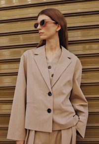 Frau mit langen braunen Haaren, trägt einen übergroßen beigen Anzug und eine Sonnenbrille, steht vor einem metallisch goldfarbenen Rollladenhintergrund.