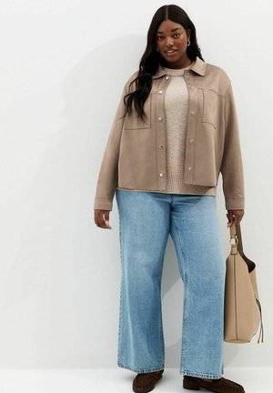 Kobieta ubrana w beżową kurtkę, kremowy sweter, szerokie jasnoniebieskie dżinsy, brązowe mokasyny, trzymająca beżową torebkę na tle białej ściany.