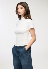 Giovane donna con capelli castani, che indossa una maglietta bianca a maniche corte aderente e jeans blu larghi, in piedi con le mani nelle tasche su uno sfondo bianco.