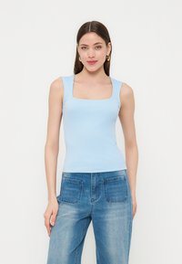 Kvinde iført en lyseblå ærmeløs top med firkantet halsudskæring og højtaljede falmede blå jeans med forlommer, stående mod en hvid baggrund.