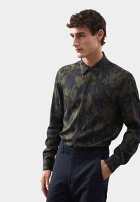 Grünes, gemustertes Langarmhemd mit einem Kragen, das abstrakte Designs zeigt, kombiniert mit navyfarbenen Hosen. Glatter Stoff und taillierte Passform.