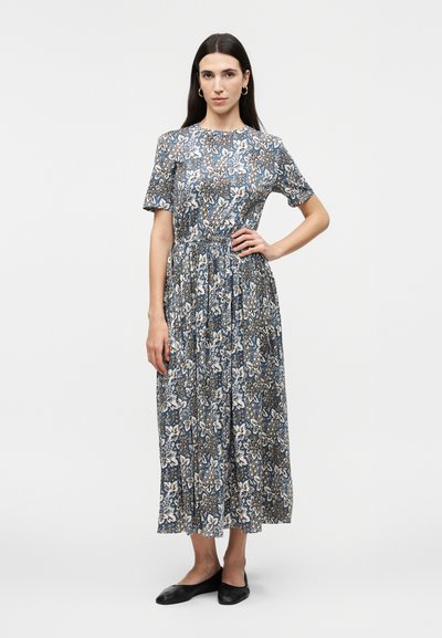 Femme aux longs cheveux foncés portant une robe midi fleurie bleue et blanche et des ballerines noires, debout avec une main sur la hanche sur un fond uni.