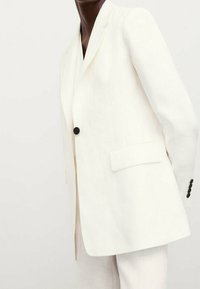 Henkilö, joka käyttää valkoista räätälöityä bleiseriä, jossa on yksi musta nappi ja läpätaskut, seisoo tasaisella vaalealla taustalla.