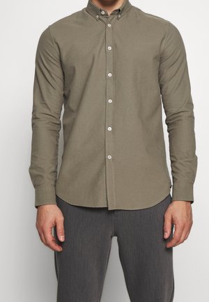 Mann trägt ein olivfarbenes, langärmliges Hemd mit Knöpfen und graue Hose, steht mit entspannten Armen an den Seiten.