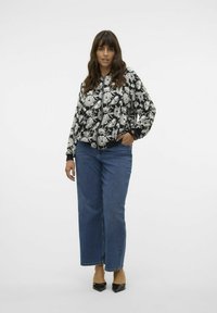 Chaqueta bomber negra con patrón floral blanco, cierre de cremallera, puños acanalados, combinada con jeans de pierna ancha azules y tacones negros de punta.