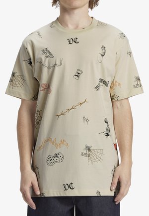 Camiseta beige con varias estampaciones gráficas, incluyendo dados, llamas y telarañas. Mangas cortas y un escote redondo.