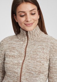 Lysebrun strikket sweater med høj krave og en frontlynlås, der har et struktureret mønster og ribbet detaljer ved kraven.