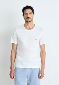 T-shirt en coton blanc avec un col rond et des manches courtes, arborant un petit logo noir "BOSS" sur la poitrine gauche. Porté avec un pantalon bleu clair.