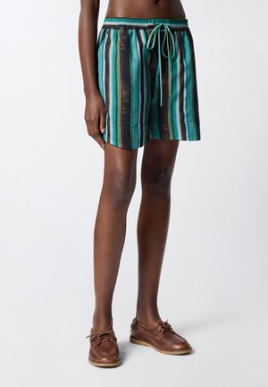 Persoon die turquoise, zwart en bruine verticale gestreepte shorts met trekkoord draagt, staand in bruine leren bootschoenen op een lichte achtergrond.