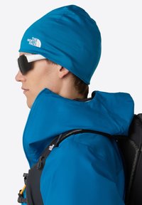 Osoba nosząca niebieską czapkę North Face, niebieską kurtkę z kapturem, czarny plecak i białe okulary sportowe, zwrócona w lewo.