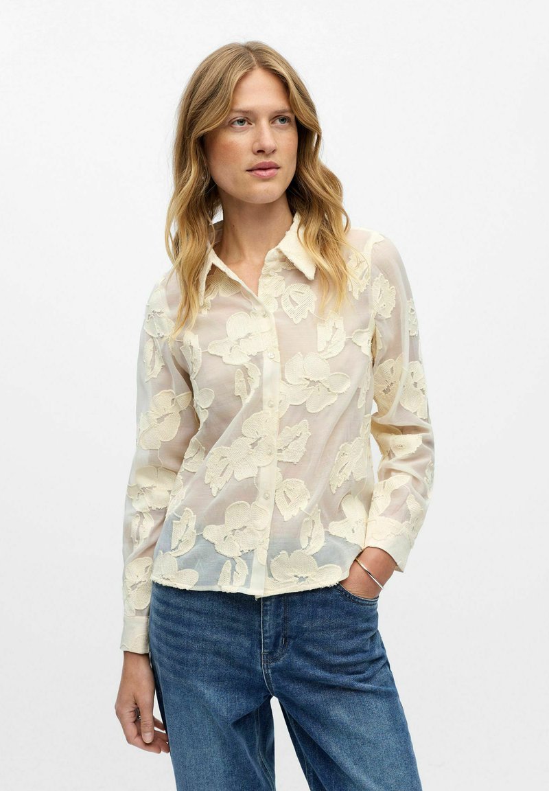 Gennemsigtig blomsterbluse i creme med en knaplukning foran, der har indviklede broderede blomstermønstre og en klassisk krave.