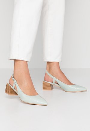 Pieds portant des chaussures slingback pointues bleu clair avec talons blocs en bois et pantalon blanc raccourci sur un fond clair uni.