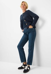 Cardigan blu navy con bottoni dorati, abbinato a jeans dritti blu scuro e sneakers nere con dettagli bianchi. Aspetto casual e aderente.