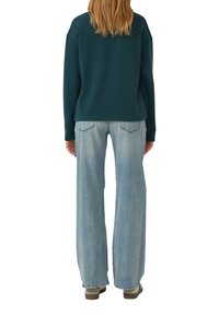 Personne avec de longs cheveux blonds portant un haut à manches longues vert foncé, un jean large bleu clair et des chaussures beiges, se tenant de dos.