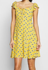 Keltainen mekko paisley-kuviolla, röyhelösateilla, neliön muotoisella pääntiellä ja solmusiimalla edessä. A-linjainen siluetti, polvipituinen.