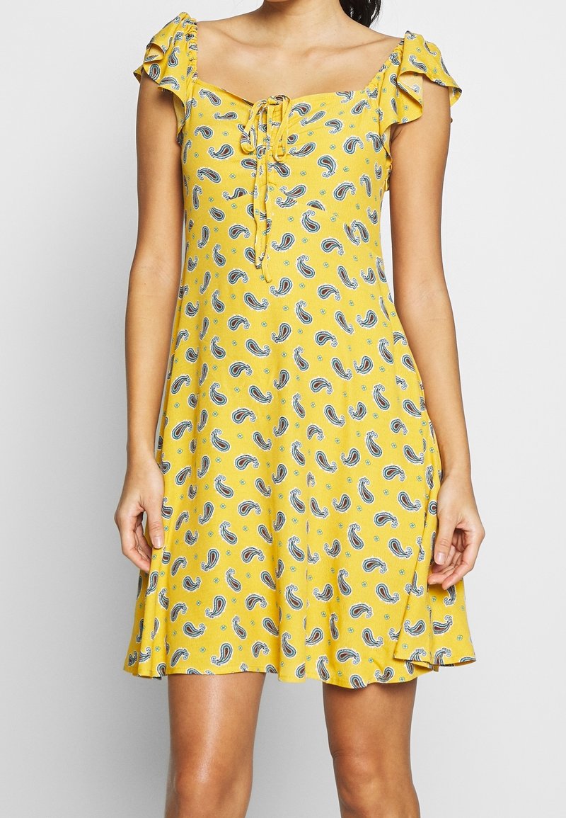 Keltainen mekko paisley-kuviolla, röyhelösateilla, neliön muotoisella pääntiellä ja solmusiimalla edessä. A-linjainen siluetti, polvipituinen.