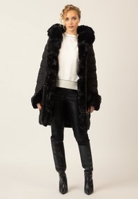 Schwarze Daunenjacke mit einem Kapuze und Manschetten aus Kunstfell, ausgestattet mit einem Reißverschluss. Getragen über einem weißen Pullover und schwarzen Lederhosen.