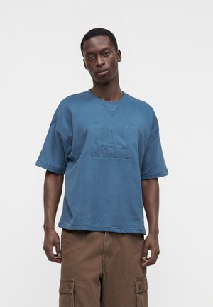 Homme portant une chemise bleue ample à manches courtes avec des lettres en relief "GAP" et un pantalon cargo marron, debout devant un fond uni.