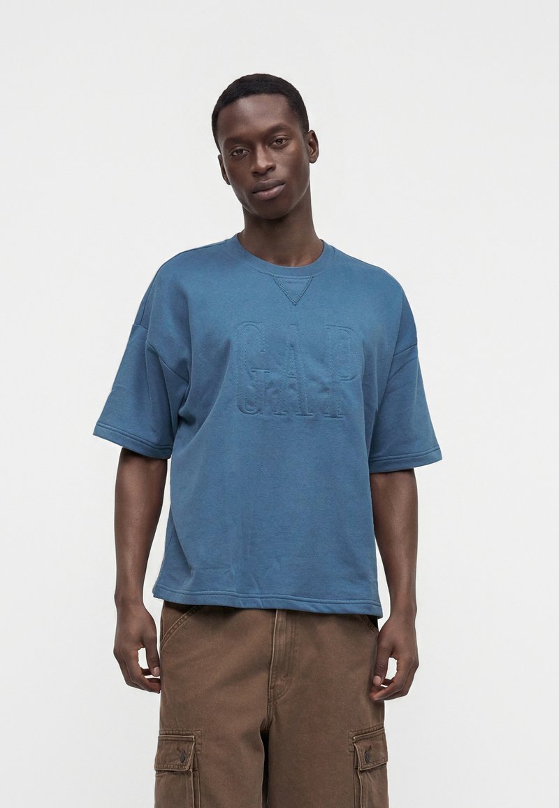Homme portant une chemise bleue ample à manches courtes avec des lettres en relief "GAP" et un pantalon cargo marron, debout devant un fond uni.