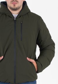 Gevoerde groene jas met capuchon, ritssluiting aan de voorkant, twee zijzakken en een klein ritszakje op de borst. Soepele textuur.