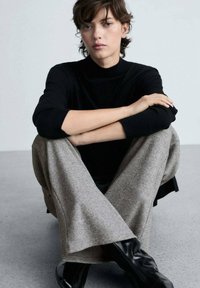 Crna turtleneck dolčevita s reljefnim rukavima, uparen s svijetlosivim hlačama širokih nogavica. Model sjedi s prekriženim nogama, noseći crne čizme do gležnja.