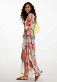 Kolorowy, wzorzysty długi kimono w odcieniach różu, niebieskiego i zielonego. Lekki materiał, otwarty przód, noszony na szortach dżinsowych i sandałach.