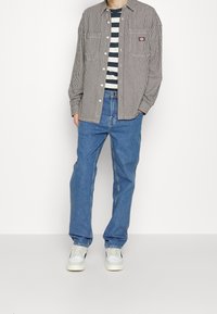 Camicia a righe beige e nera sopra una maglietta a righe blu navy; abbinata a jeans in denim blu e sneaker bianche.