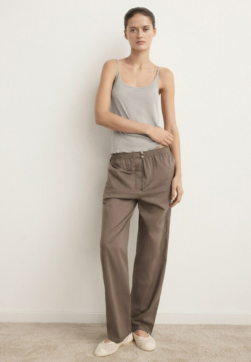 Femme debout contre un mur blanc, portant un débardeur gris, un pantalon marron ample et des chaussures blanches tissées sans lacets.