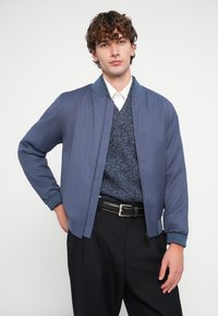 Μπλε μπουφάν τύπου bomber με φερμουάρ μπροστά, ριμπ μανσέτες και γιακά, φορεμένο πάνω από ένα μπλε πλεκτό πουλόβερ με υφή και λευκό πουκάμισο, συνδυασμένο με μαύρο παντελόνι.