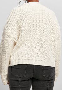 Person som bär en krämfärgad grovstickad tröja och svarta jeans, sedd bakifrån mot en enfärgad ljus bakgrund.