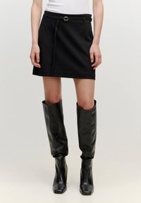 Schwarzer Rock über dem Knie mit einem Gürtel-Detail, kombiniert mit hohen schwarzen Lederstiefeln. Die glatte Textur und das taillierte Design betonen die Silhouette.