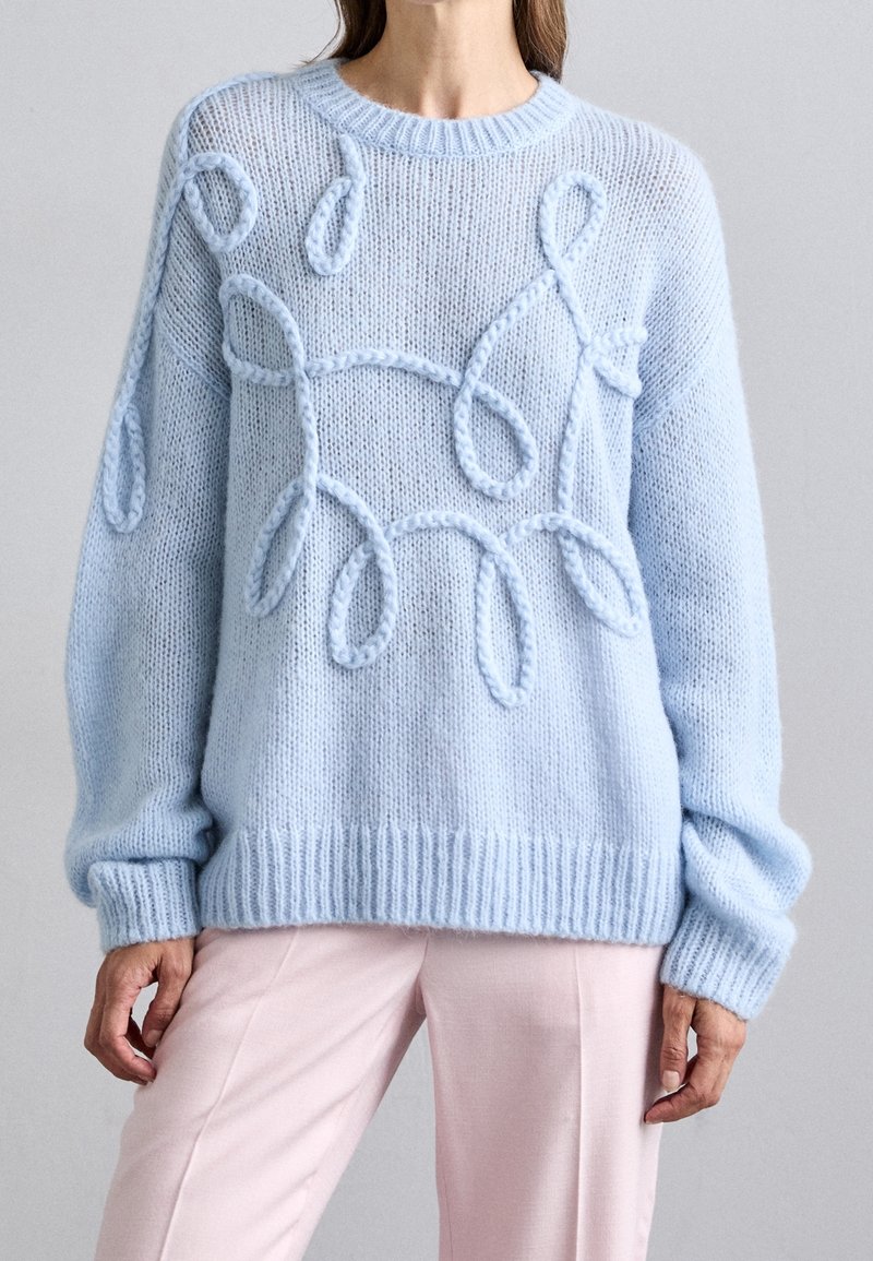 Femme portant un pull tricoté bleu clair avec des motifs en boucles en relief, associé à un pantalon rose clair sur un fond uni.