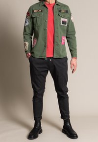 Man som bär en grön jacka med lappar över en röd skjorta, svarta smala byxor och svarta läderstövlar, står med händerna i fickorna.