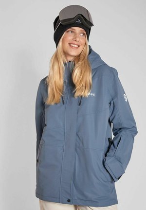 Kvinde iført en blå vinterjakke, sort hue og skibriller på panden, smilende og kigger til siden.