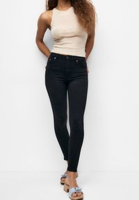 Pantalones vaqueros ajustados negros de cintura alta, combinados con una camiseta sin mangas de color beige claro y sandalias azules tipo deslizamiento con un detalle de lazo.
