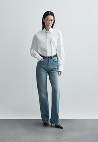 Λευκό πουκάμισο με κουμπιά και μακριά μανίκια, συνδυασμένο με ανοιχτό μπλε ψηλόμεσα τζιν και μαύρες μυτερές γόβες. Απλός σχεδιασμός, καλοκαιρινό στυλ.