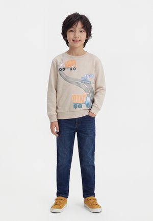 Dreng står iført beige sweatshirt med byggeplads lastbiler, blå jeans og sennepsgule sneakers mod hvid baggrund.