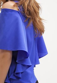 Blå blus med off-the-shoulder-design, med volangärmar och en slät tygtextur. Detaljer inkluderar en subtil metallaccent.