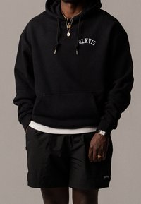 Sweat à capuche noir avec logo "BLKVIS", poche kangourou, et ourlet côtelé blanc, assorti à un short noir. Détails supplémentaires incluant des colliers dorés et une montre.