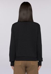 Czarny sweatshirt z długimi rękawami i ściągaczami, z okrągłym hemem i miękką, gładką teksturą. Noszony z brązowymi spodniami.