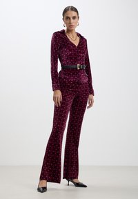 Tutina in velluto borgogna con un motivo geometrico. Presenta un colletto, maniche lunghe e una cintura nera in vita. Design a gamba svasata.