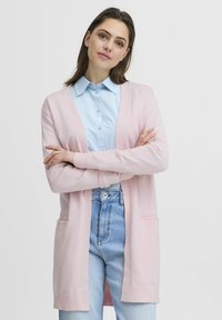 Cardigan rosa chiaro con maniche lunghe e apertura frontale. Presenta tasche laterali e polsini a coste. Indossato sopra una camicia colletto azzurra.