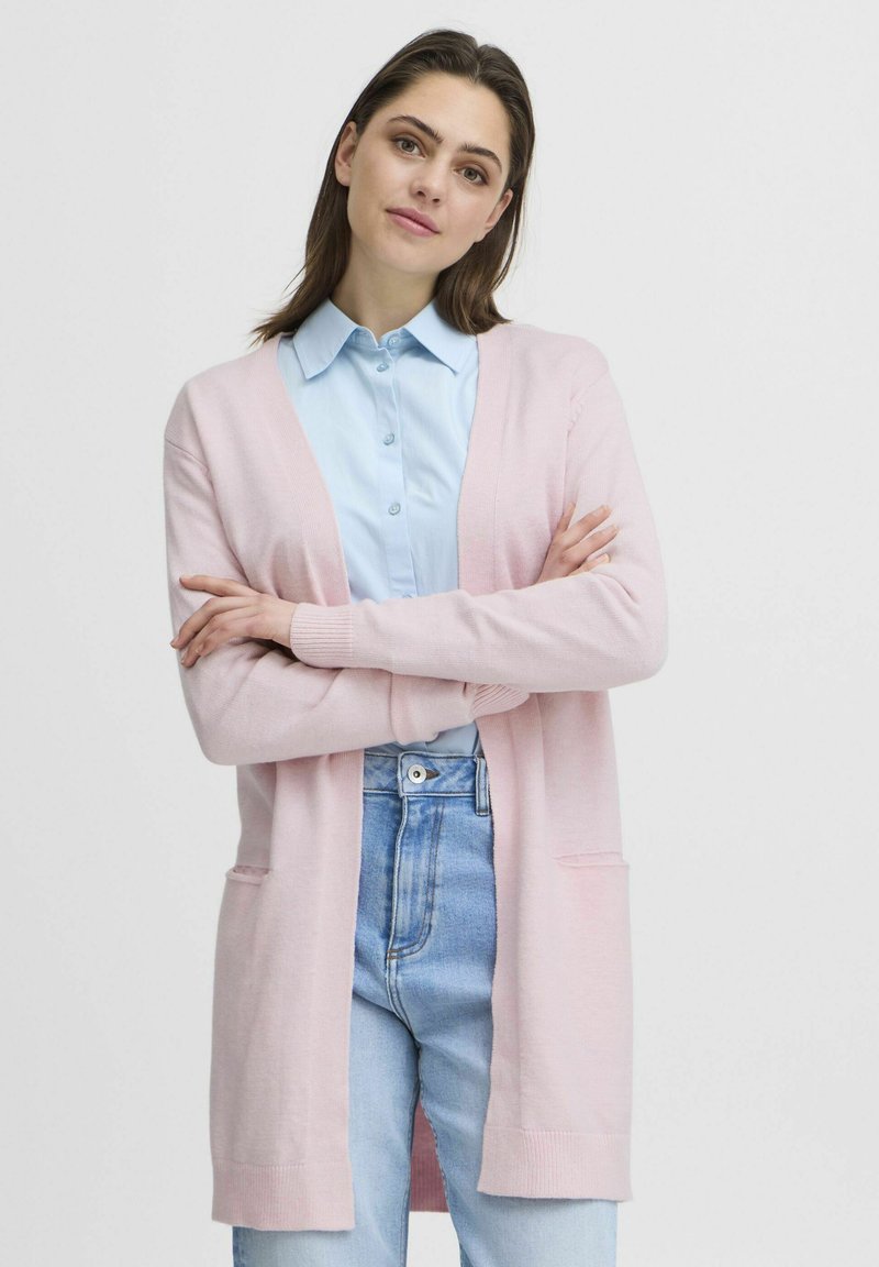 Cardigan rosa chiaro con maniche lunghe e apertura frontale. Presenta tasche laterali e polsini a coste. Indossato sopra una camicia colletto azzurra.